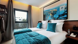 Camera d'albergo con due letti, vista sulla città, un quadro appeso e un comodino con un telefono e una bottiglia d'acqua.