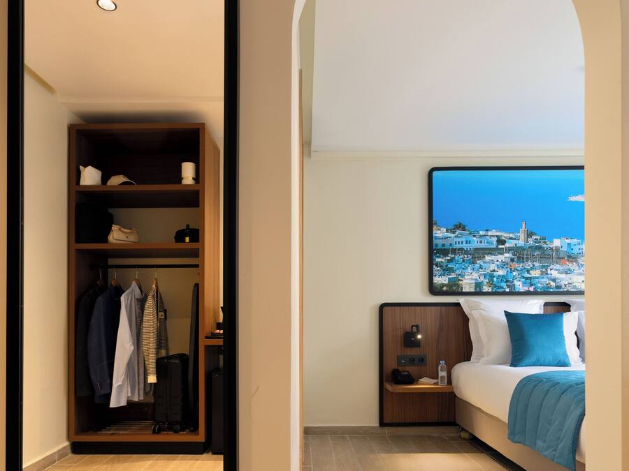 Een hotelkamer met een kledingkast, een bed met uitzicht op een kustplaats, een nachtkastje en een aan de muur bevestigde televisie.
