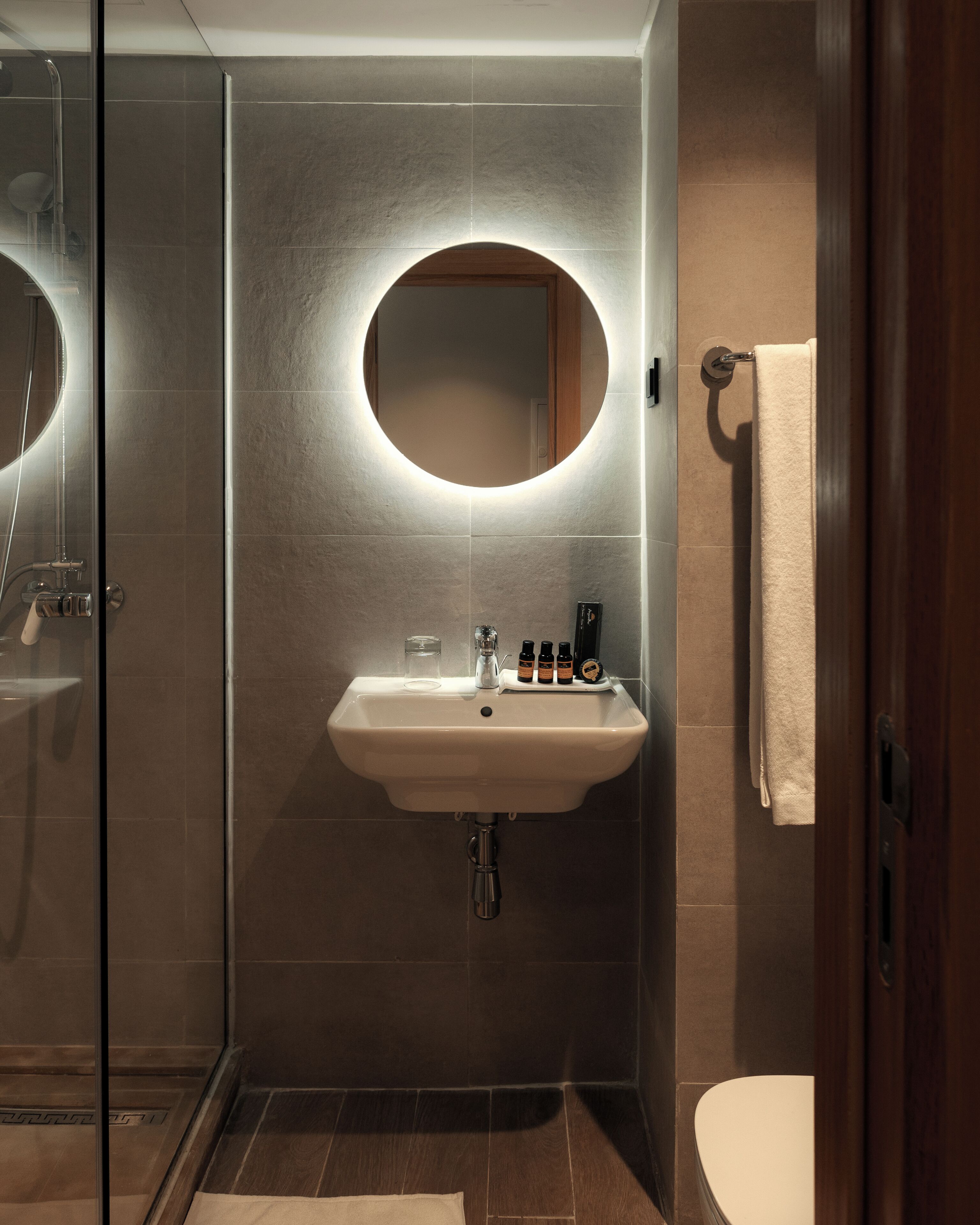 A modern bathroom with a glass-enclosed shower, a round mirror with underglow lighting, a white sink with toiletries, and a wooden door.