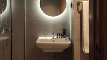 A modern bathroom with a glass-enclosed shower, a round mirror with underglow lighting, a white sink with toiletries, and a wooden door.