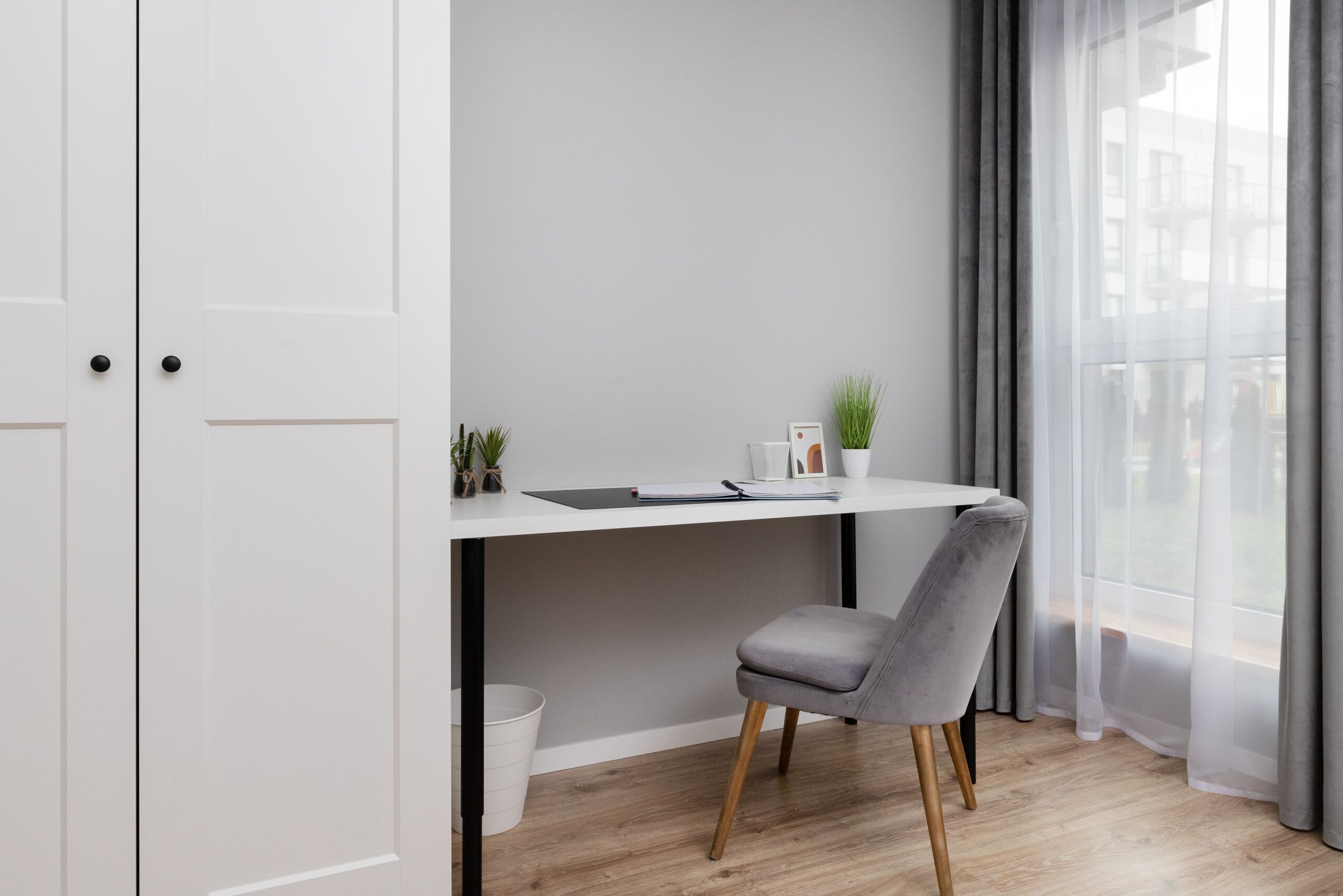 Una habitación moderna con un escritorio blanco, una silla gris, una planta pequeña y una ventana con cortinas grises.