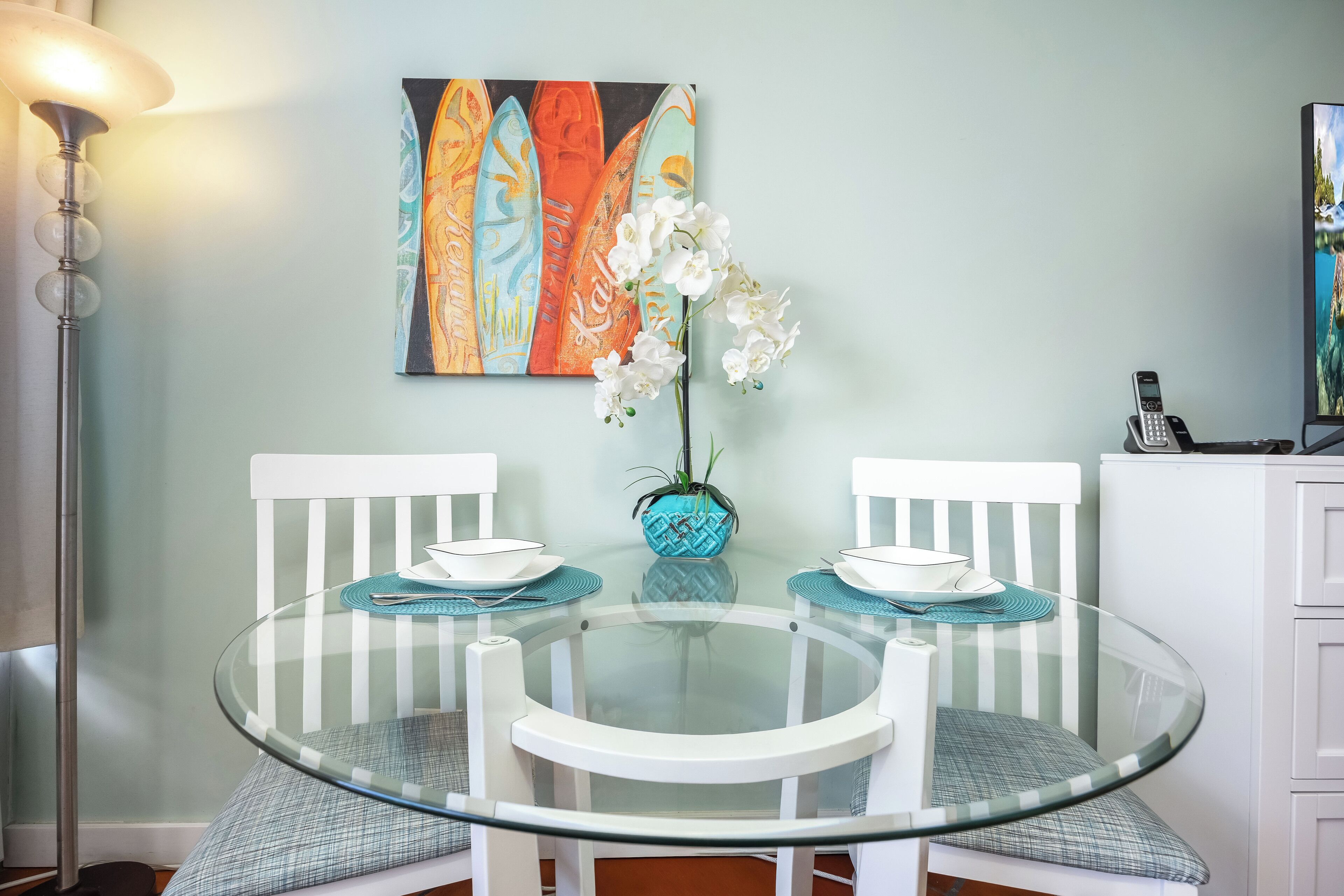 A round glass table with white chairs, a vase with white flowers, and a colorful painting on the wall.