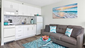A modern kitchen with white cabinets, a microwave, and a stove. There's a gray sofa with blue pillows and a painting of a beach scene on the wall. - "Bahama" Studio w/ FREE Parking & Full Kitchen (Honolulu)