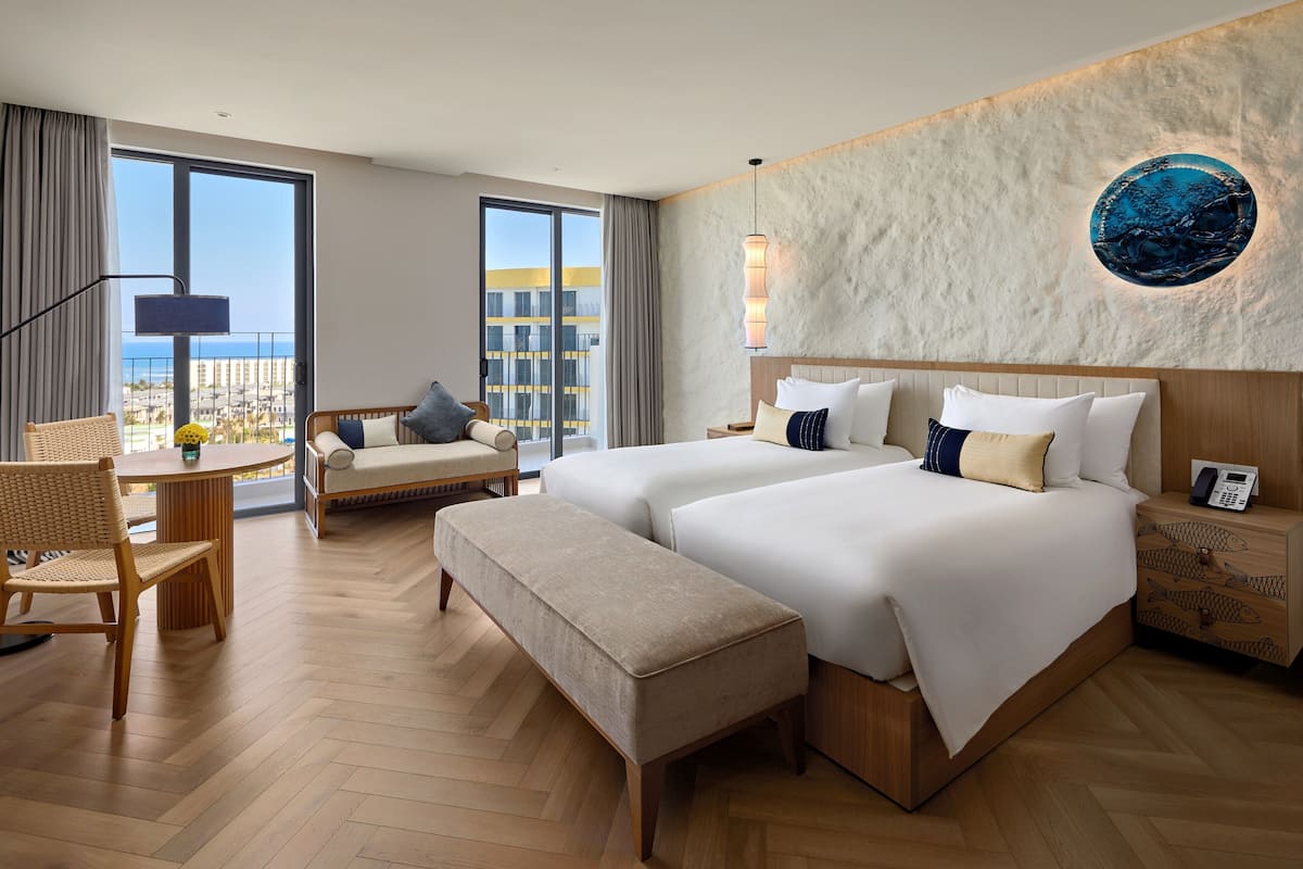 A modern hotel room with two beds, a sitting area with a table and chairs, a large window with a view, and a decorative wall piece.