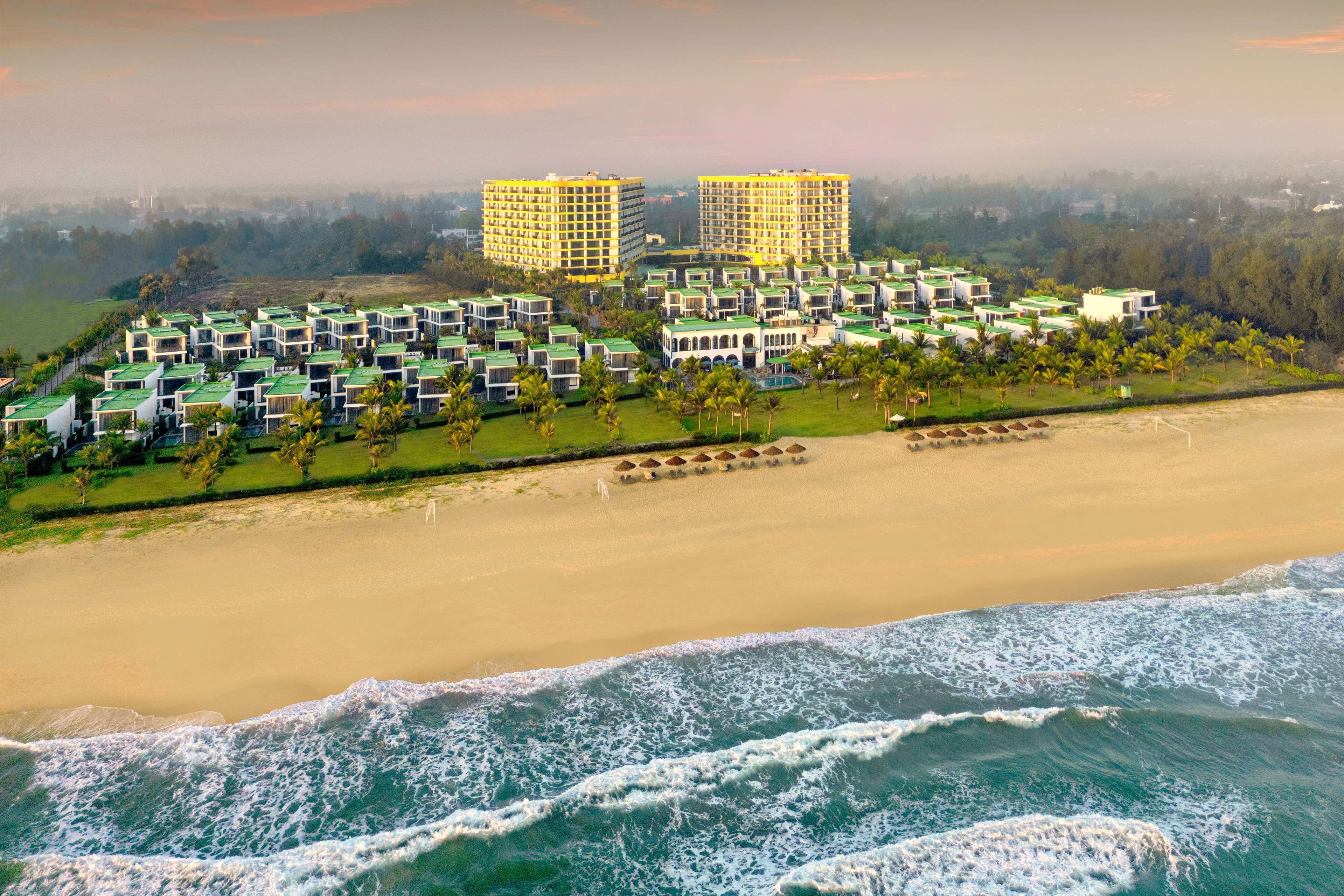 A coastal resort with multiple bungalow-style accommodations, a beachfront, and two prominent high-rise buildings in the background.