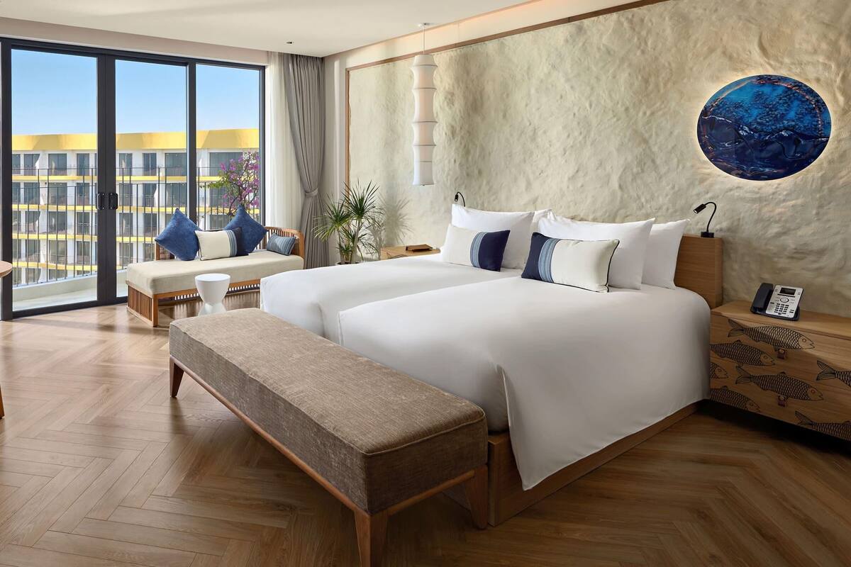 Una habitación de hotel moderna con una cama grande, un banco, una mesita de noche con teléfono y vistas al edificio.