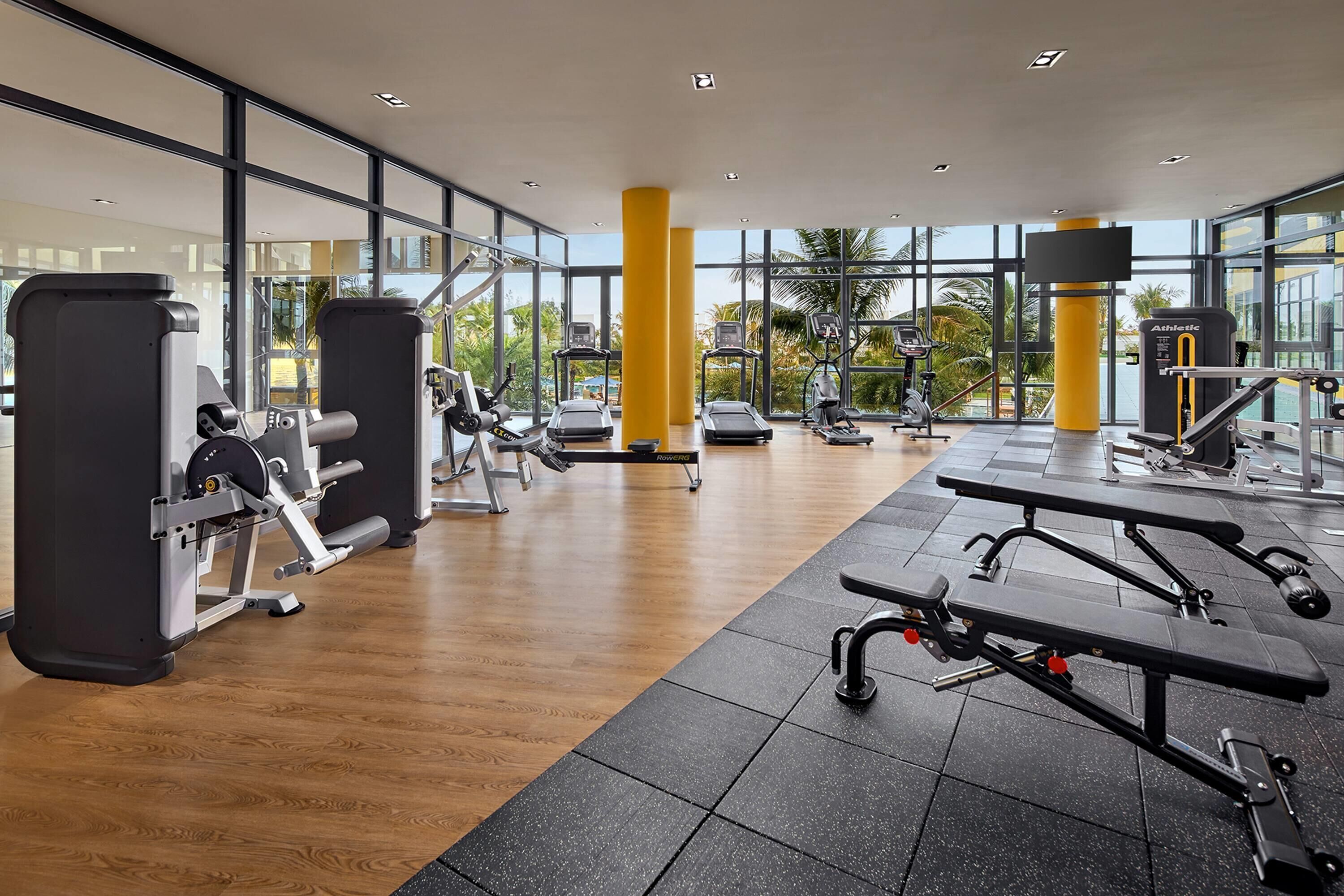 A gym with various exercise equipment, large windows, and a wooden floor.