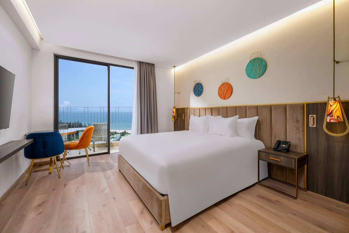 A modern hotel room with a large bed, a desk with two chairs, a nightstand, and a view of the sea through a large window.