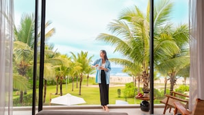 A room with a large window overlooking a beach and palm trees. - Wyndham Royal Beachfront Resort & Spa Hoi An Danang (Da Nang)