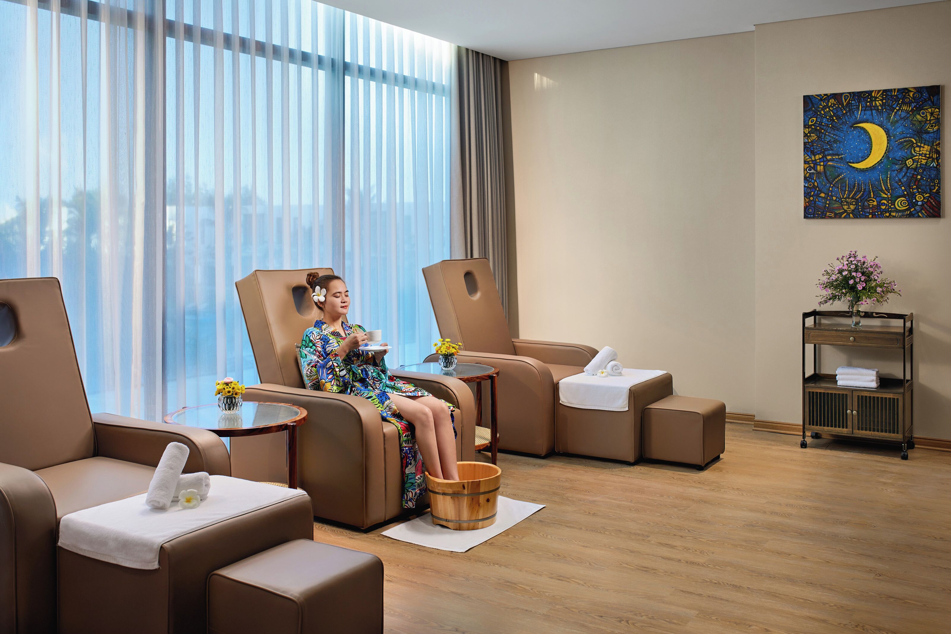 A modern hotel room with a large window, a massage chair, a small table with a vase of flowers, and a wooden bucket.