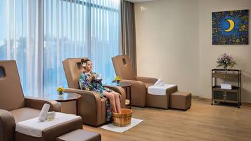 A modern hotel room with a large window, a massage chair, a small table with a vase of flowers, and a wooden bucket.