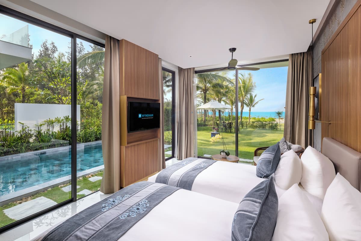 A modern hotel room with a large bed, a TV, and a view of a swimming pool and tropical landscape.