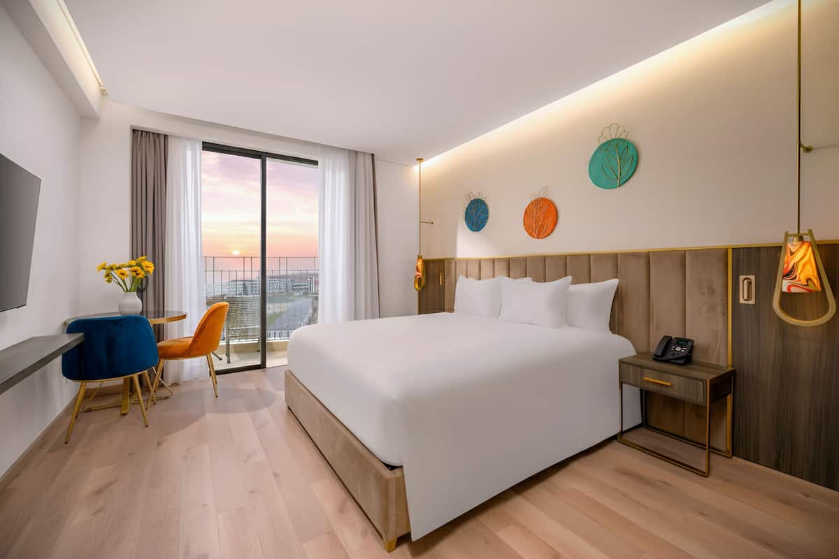 A modern hotel room with a large bed, a desk with a chair, a small table with a vase of flowers, and a city view through the window.