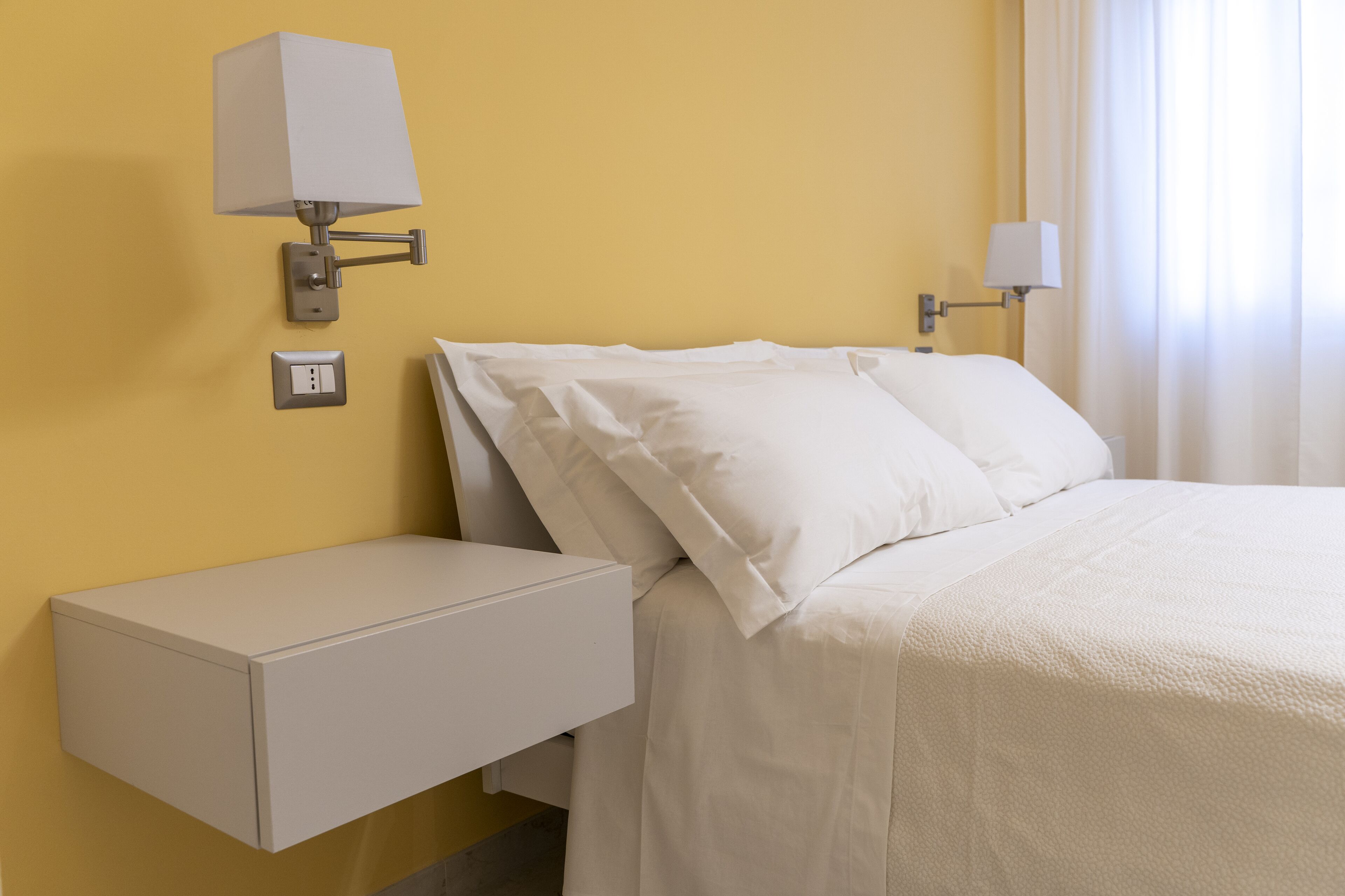 A hotel room with a bed, a nightstand, a wall-mounted lamp, and a power outlet.