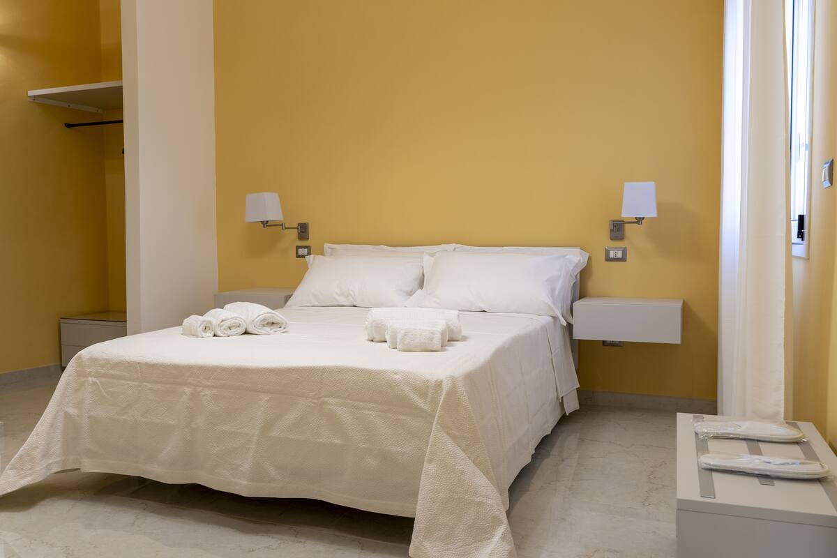 A neatly made bed with white linens and towels, a bedside table with a lamp, and a wall-mounted shelf.