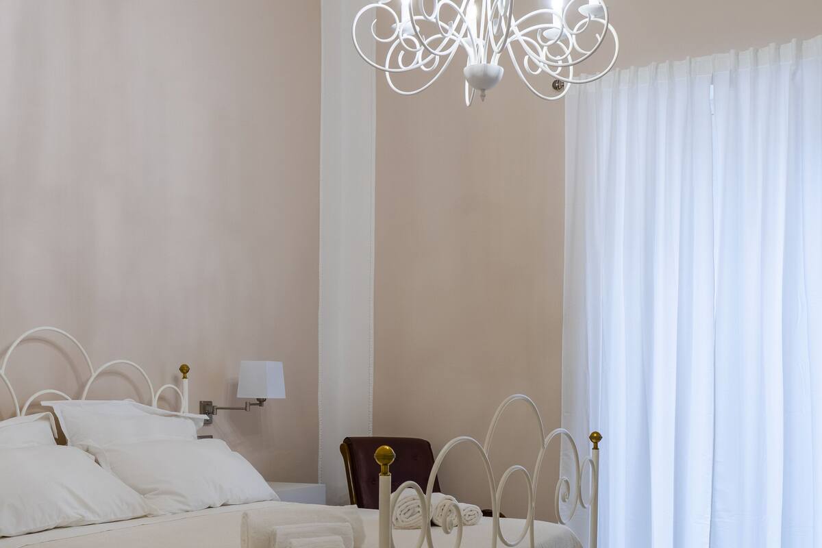 A neatly made bed with a white headboard, a bedside table with a lamp, and a chandelier.