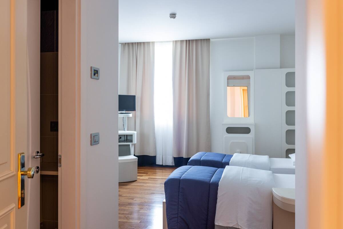 A hotel room with a wooden floor, two beds with blue bedding, a TV, a small window with curtains, and a white bathroom with a mirror and sink.