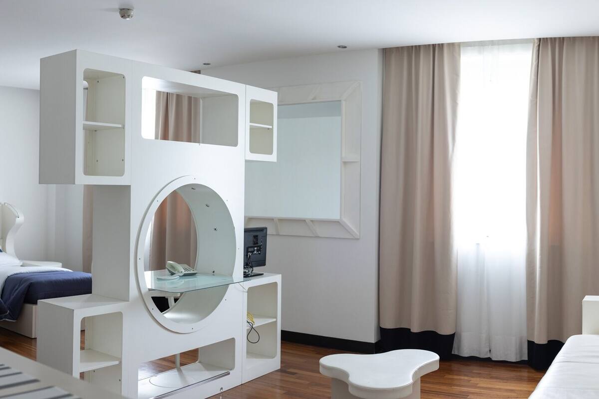 A modern hotel room with a bed, a desk with a mirror, a small table, and a large window with curtains.