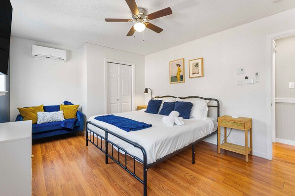 A bedroom with a bed, a ceiling fan, an air conditioner, a wooden floor, a blue sofa, and a small wooden side table.