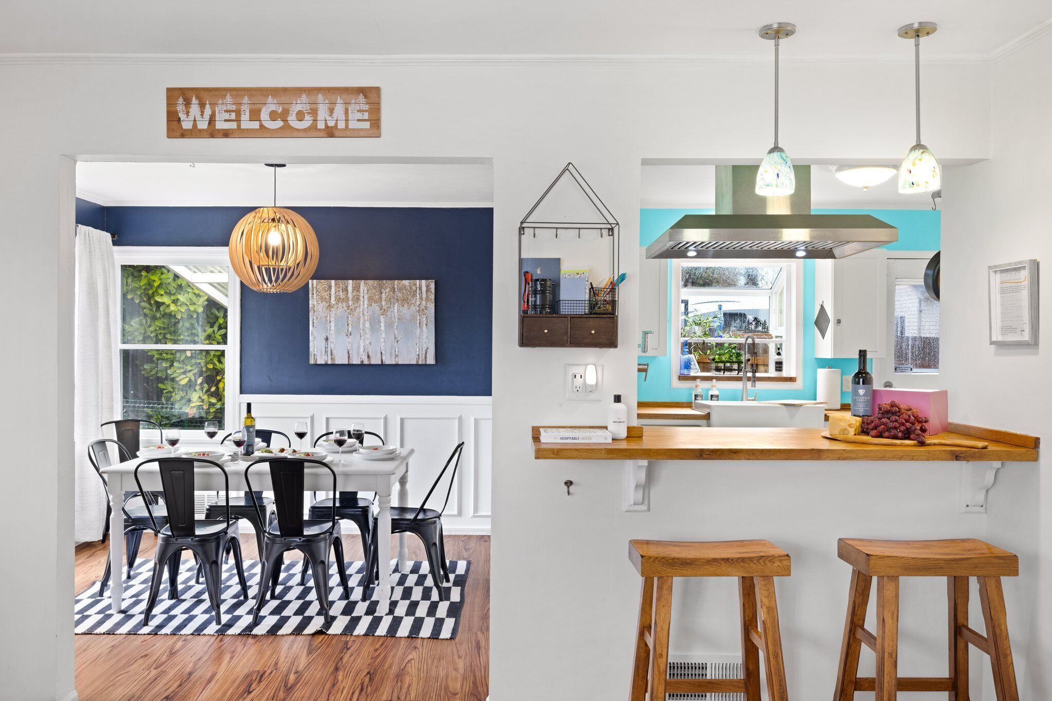 A modern kitchen with a bar area, a dining table with chairs, and a 'WELCOME' sign above the entrance.