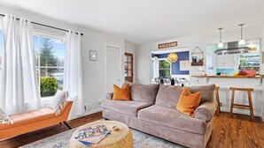 A living room with a sofa, ottoman, and a rug. There is a kitchen area with a 'Welcome' sign and a hanging light fixture. - Hot Tub! Pets! Green  Clean-Walking Distance to Proctor District (Tacoma)