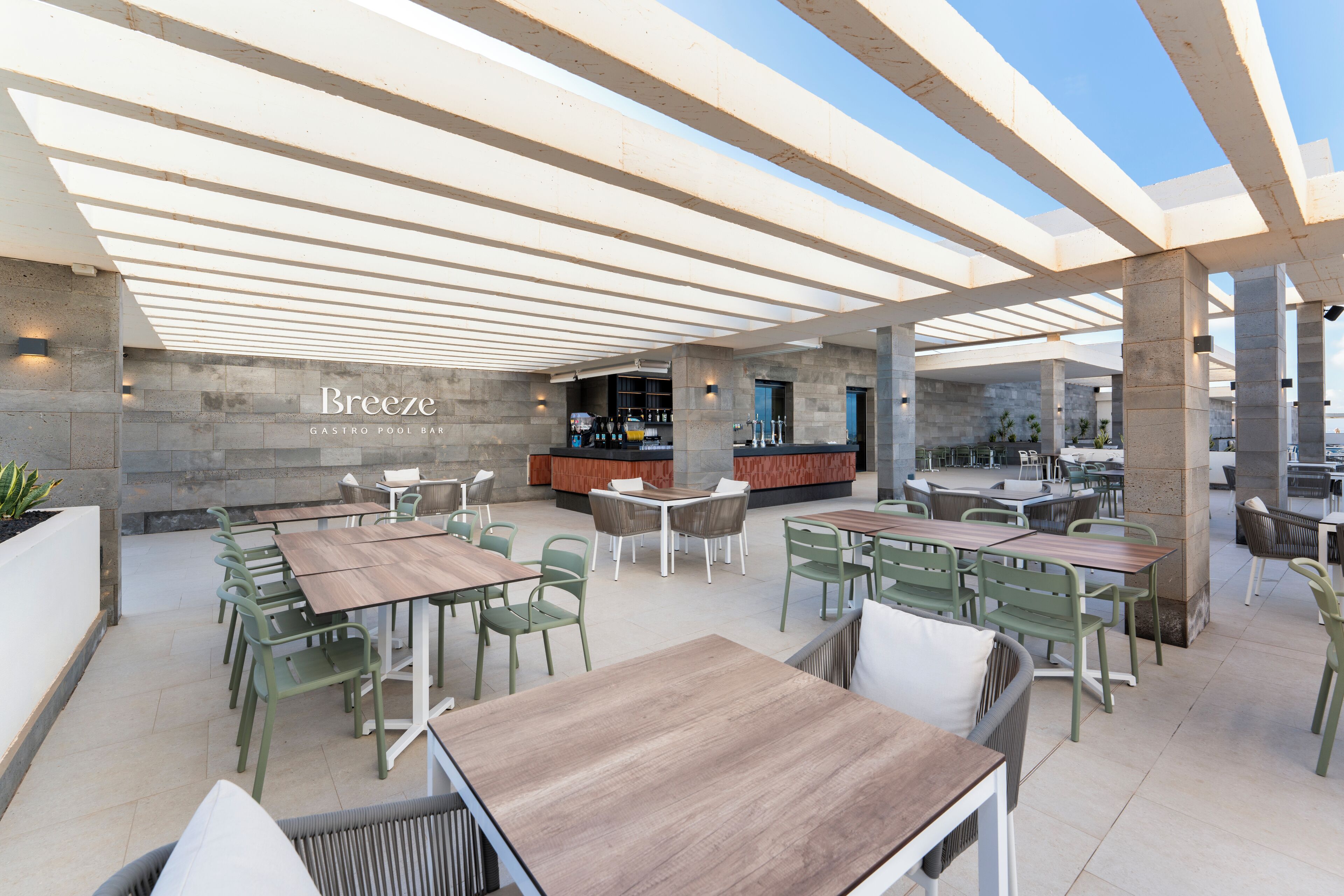 Un espace de restauration en plein air avec plusieurs tables en bois et des chaises vertes, un bar à l’arrière-plan et un mur de pierre sur lequel est inscrit le nom « Breeze ».