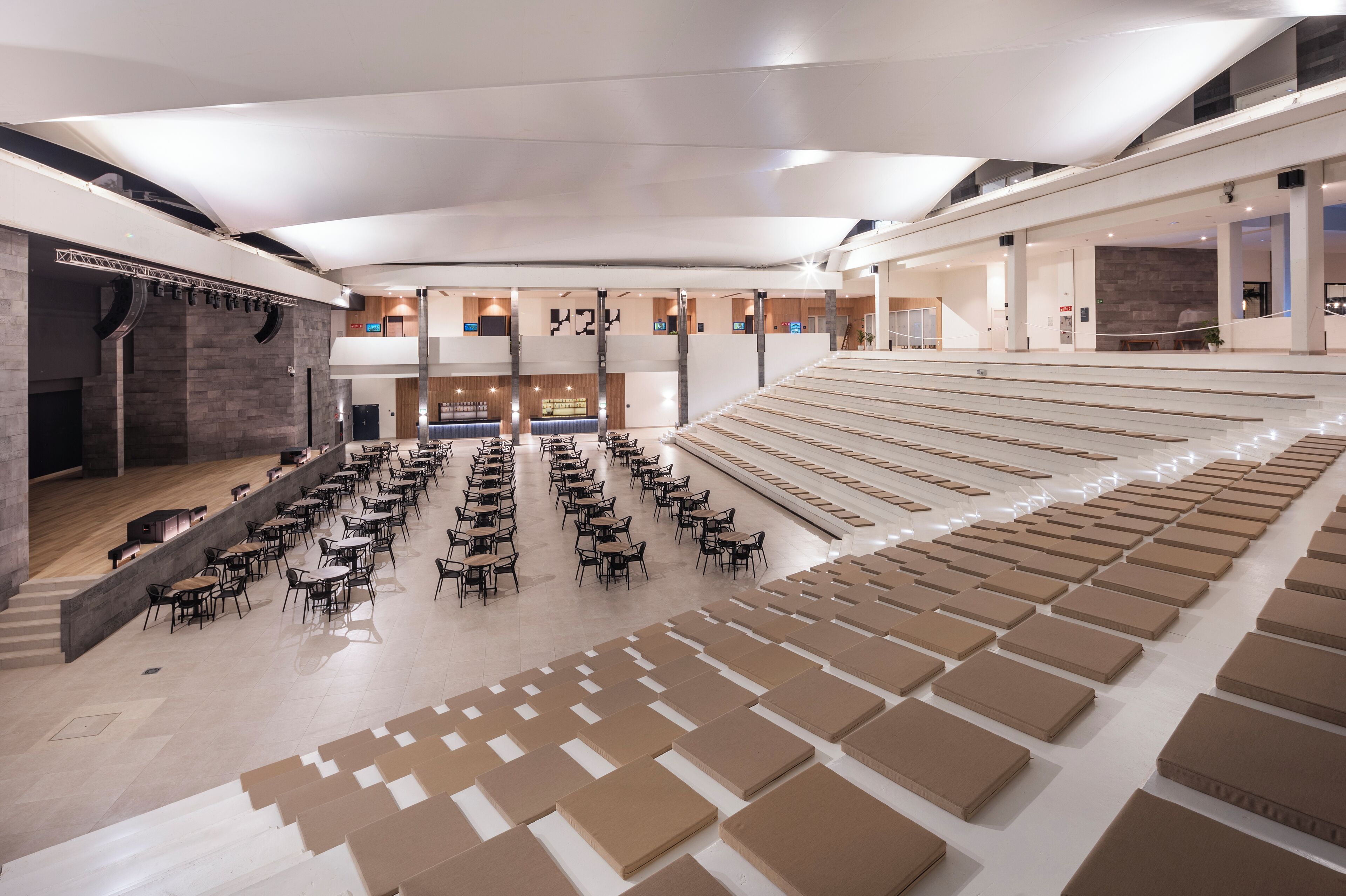Un espacio interior amplio con asientos escalonados y mesas, un escenario y un diseño arquitectónico moderno.