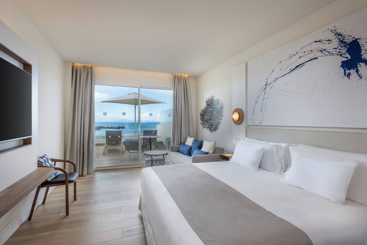 Ein Hotelzimmer mit einem großen Bett, einer Sitzecke inklusive Sofa und Sesseln, einem Flachbildfernseher und Meerblick durch eine Glastür.