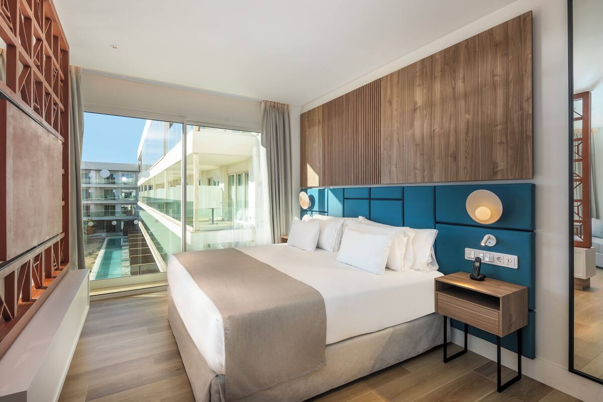 Ein modernes Hotelzimmer mit einem großen Bett, einem Nachttisch mit Telefon, Blick auf einen Pool und andere Zimmer sowie einer Wand mit Holzelementen.