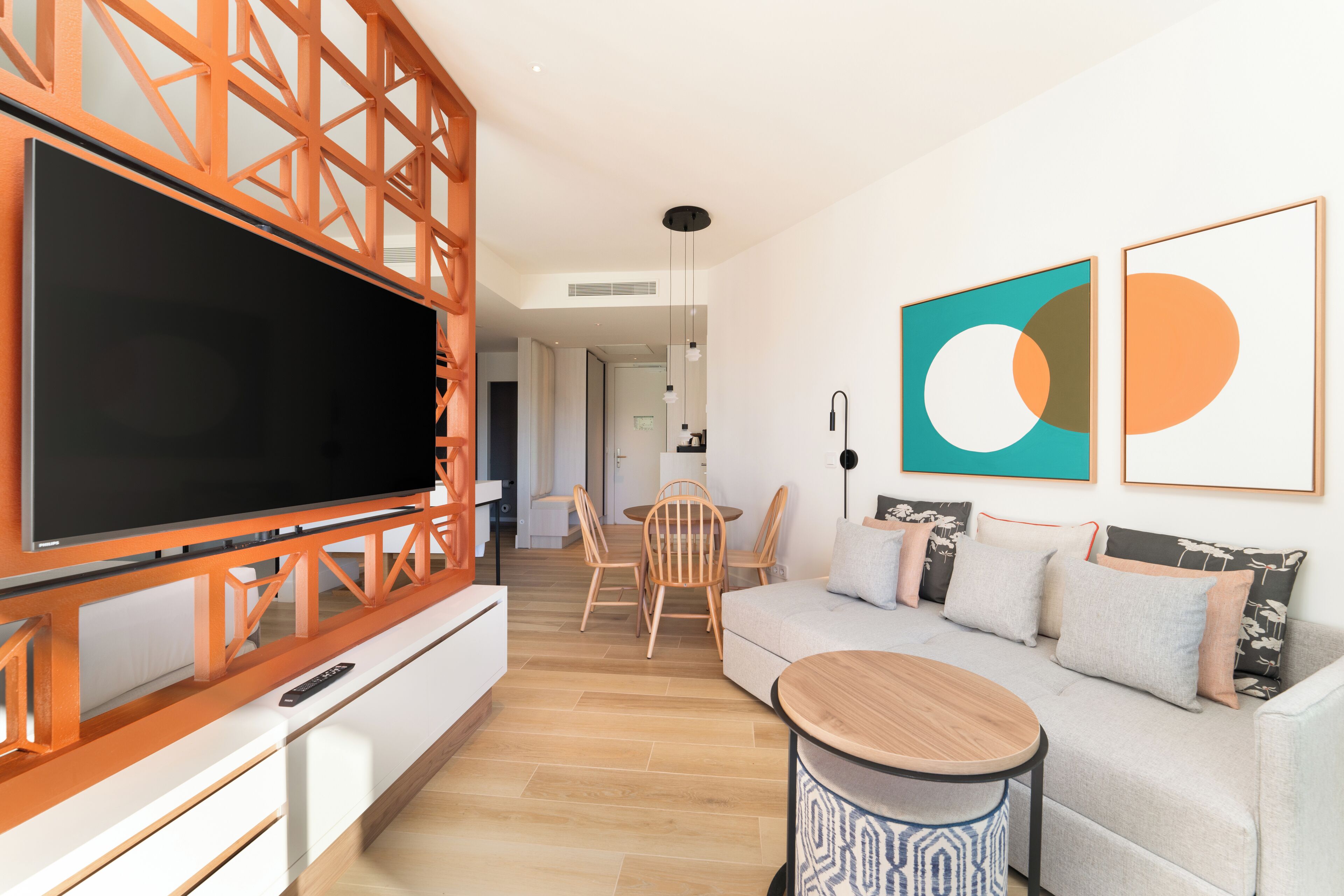 A modern living room with a large flat-screen TV, a wooden TV stand, a round wooden coffee table, a grey sofa with cushions, and two abstract wall art pieces.