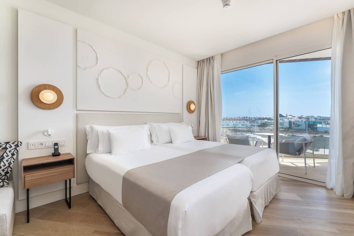 Ein modernes Hotelzimmer mit einem großen Bett, einem Nachttisch mit Lampe, Blick auf die Stadt durch große Fenster und einem Balkon mit Tisch und Stühlen.