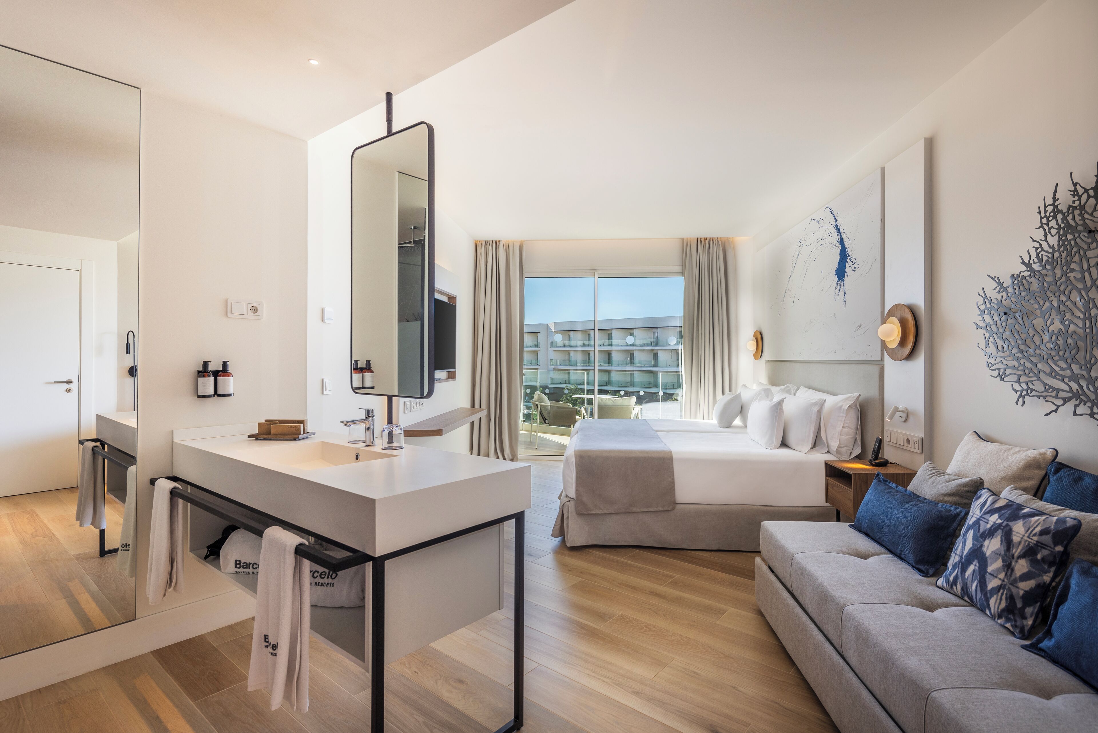 A modern hotel room with a large bed, a sofa, a bathroom with a mirror, and a balcony with a view of the city.