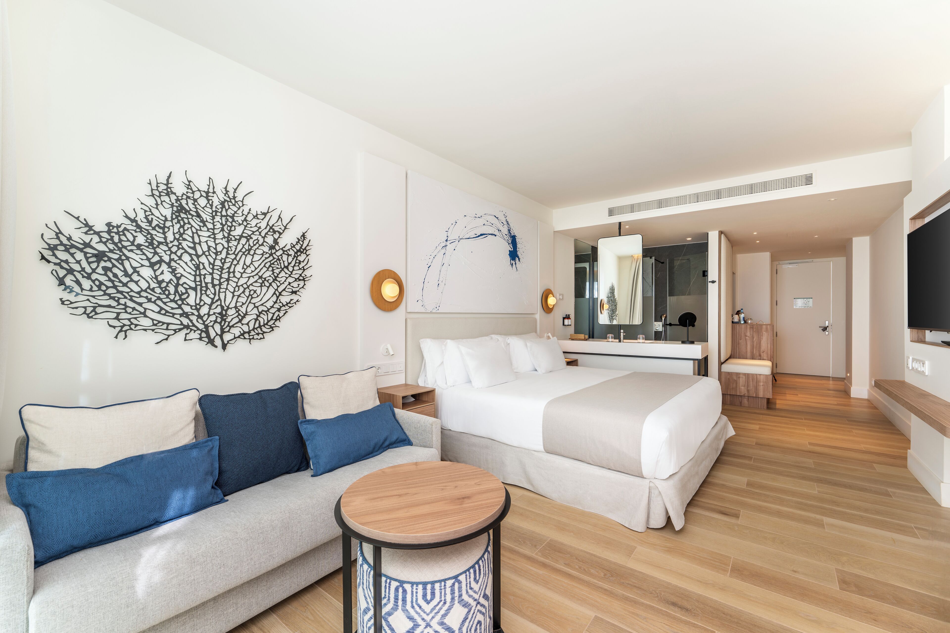 A modern hotel room with a sofa, a round wooden table, a bed, a large mirror, and a wall art piece.