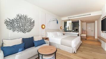 A modern hotel room with a sofa, a round wooden table, a bed, a large mirror, and a wall art piece.