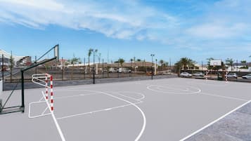 An outdoor basketball court with a goal, surrounded by a fence and palm trees.