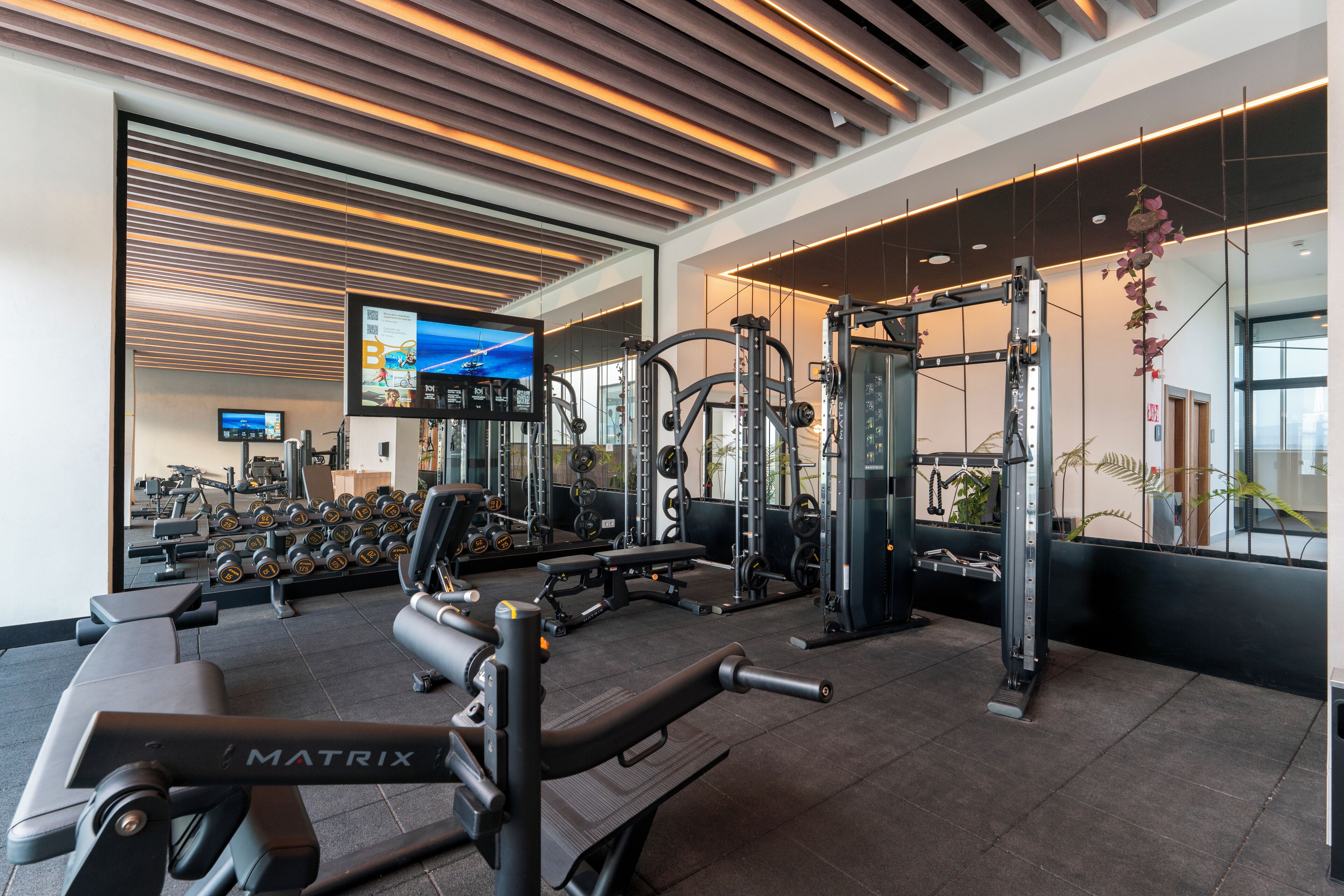 Un gimnasio moderno con diversos equipos de ejercicio, una pantalla de televisión y un espejo grande.