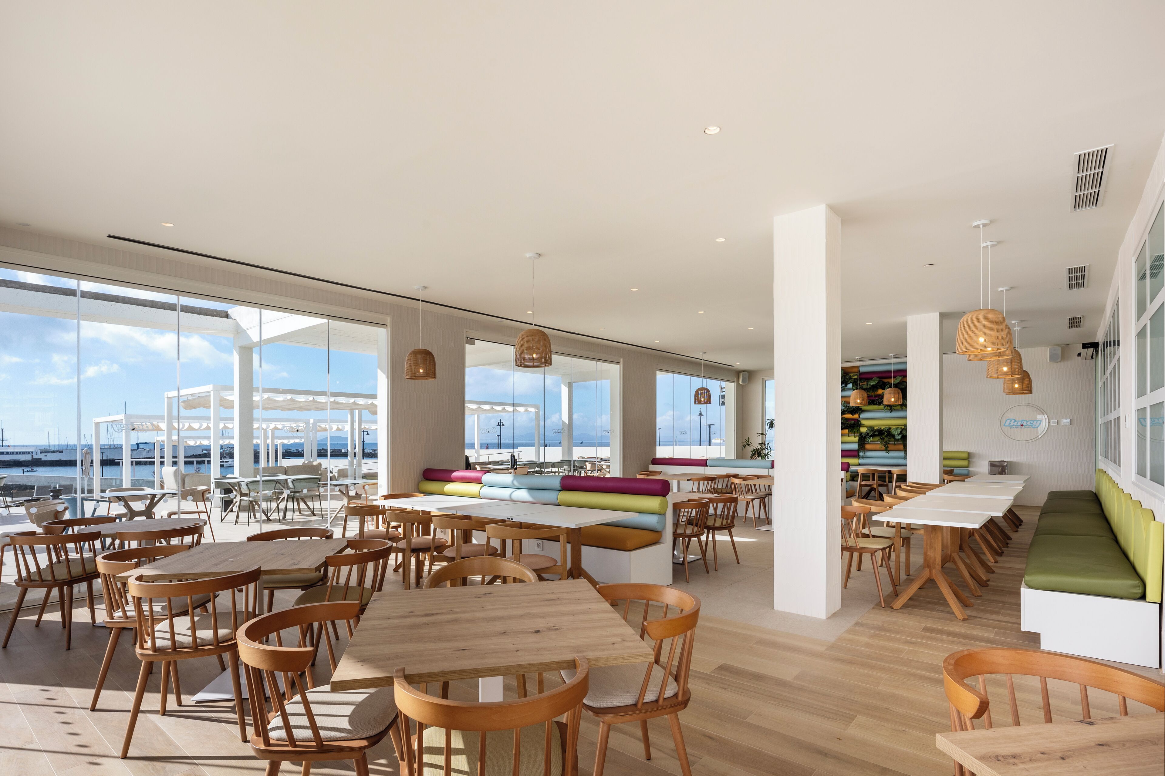 A spacious indoor dining area with wooden tables and chairs, large windows offering views of the sea, and a colorful seating area.