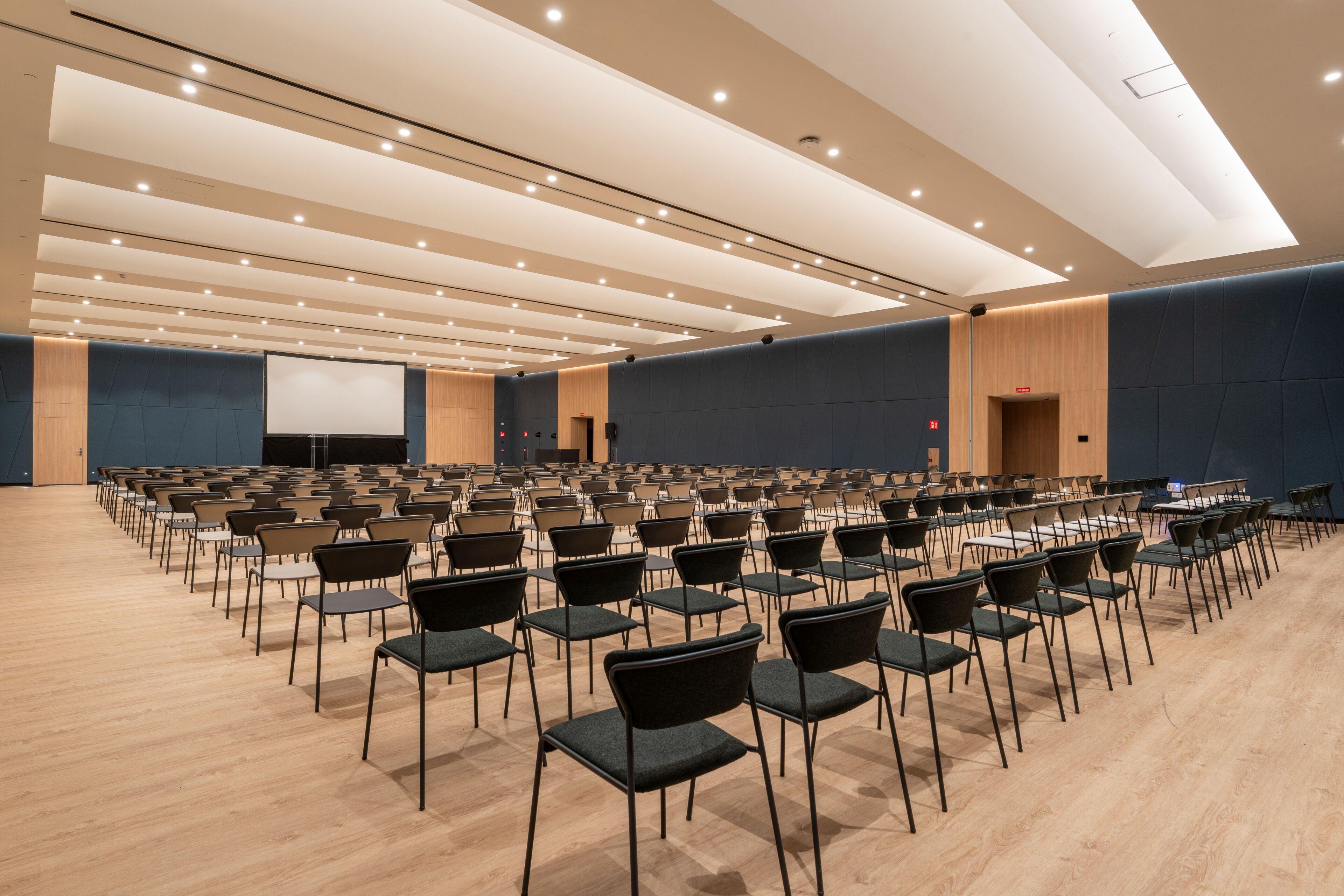 A large conference room with rows of chairs, a projector screen, and a wooden panel wall.