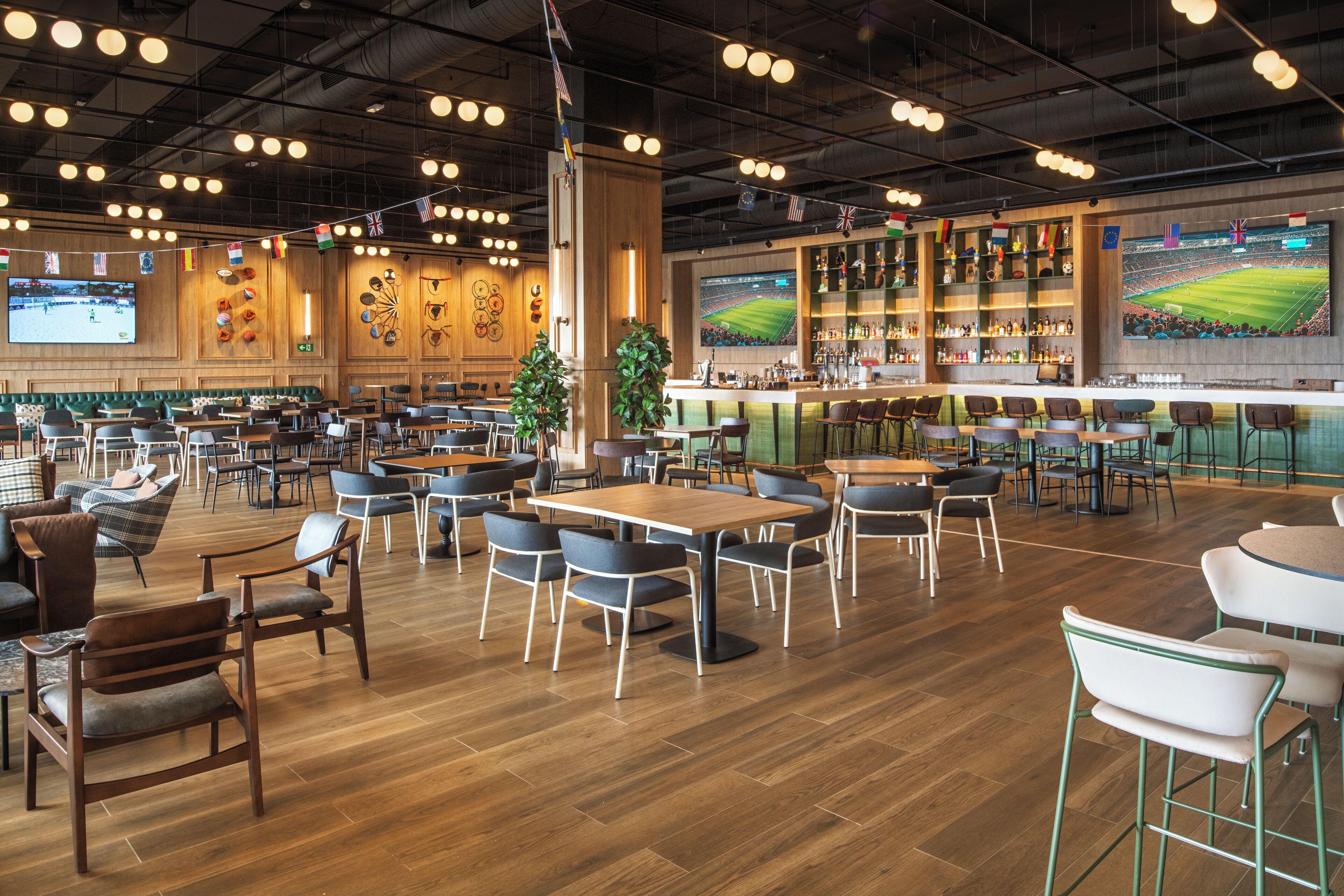 A spacious indoor dining area with multiple tables and chairs, a bar area with a large screen displaying a sports event, and various flags hanging from the ceiling.