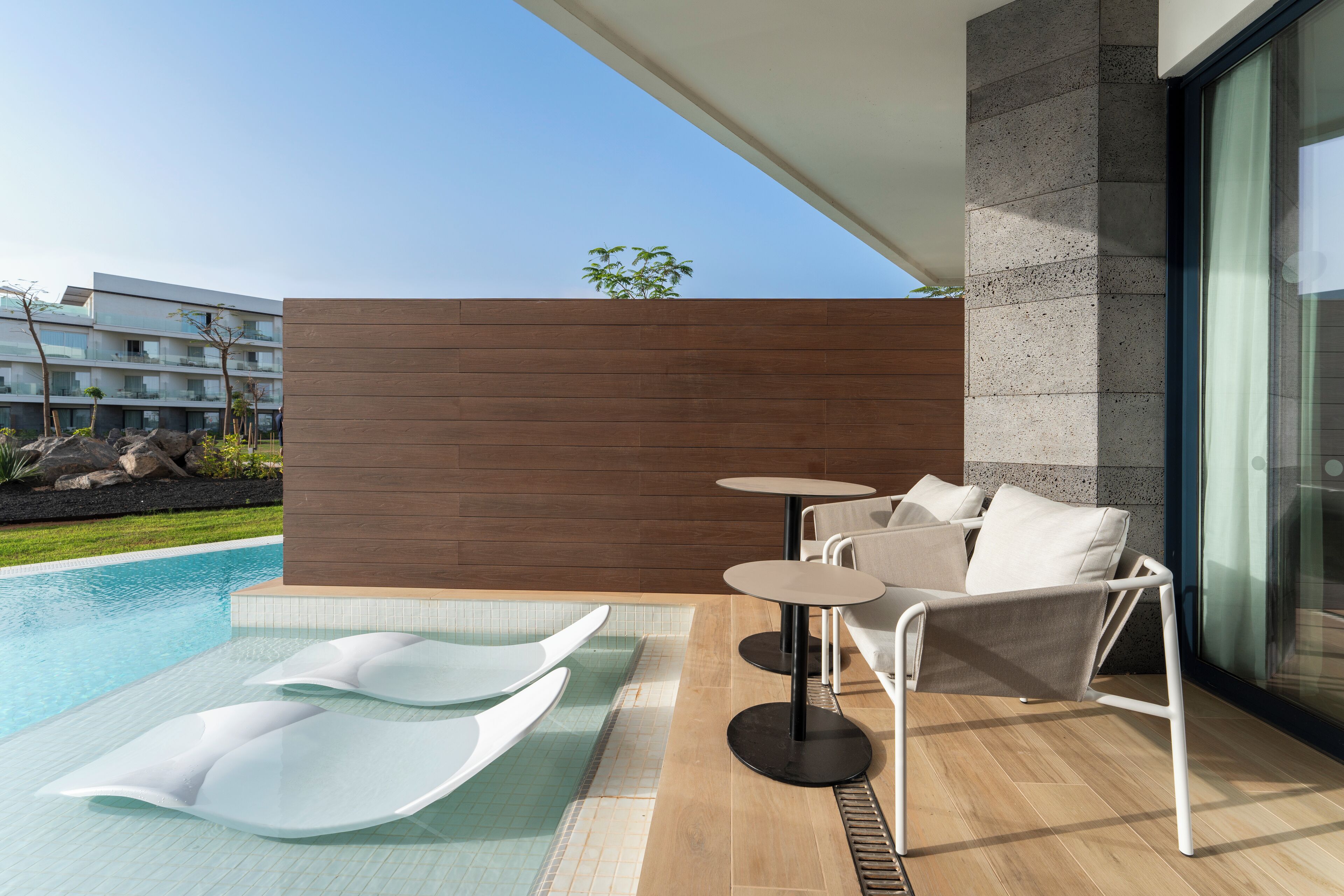 A modern outdoor pool area with lounge chairs and a table, a wooden wall, and a view of a building in the background.
