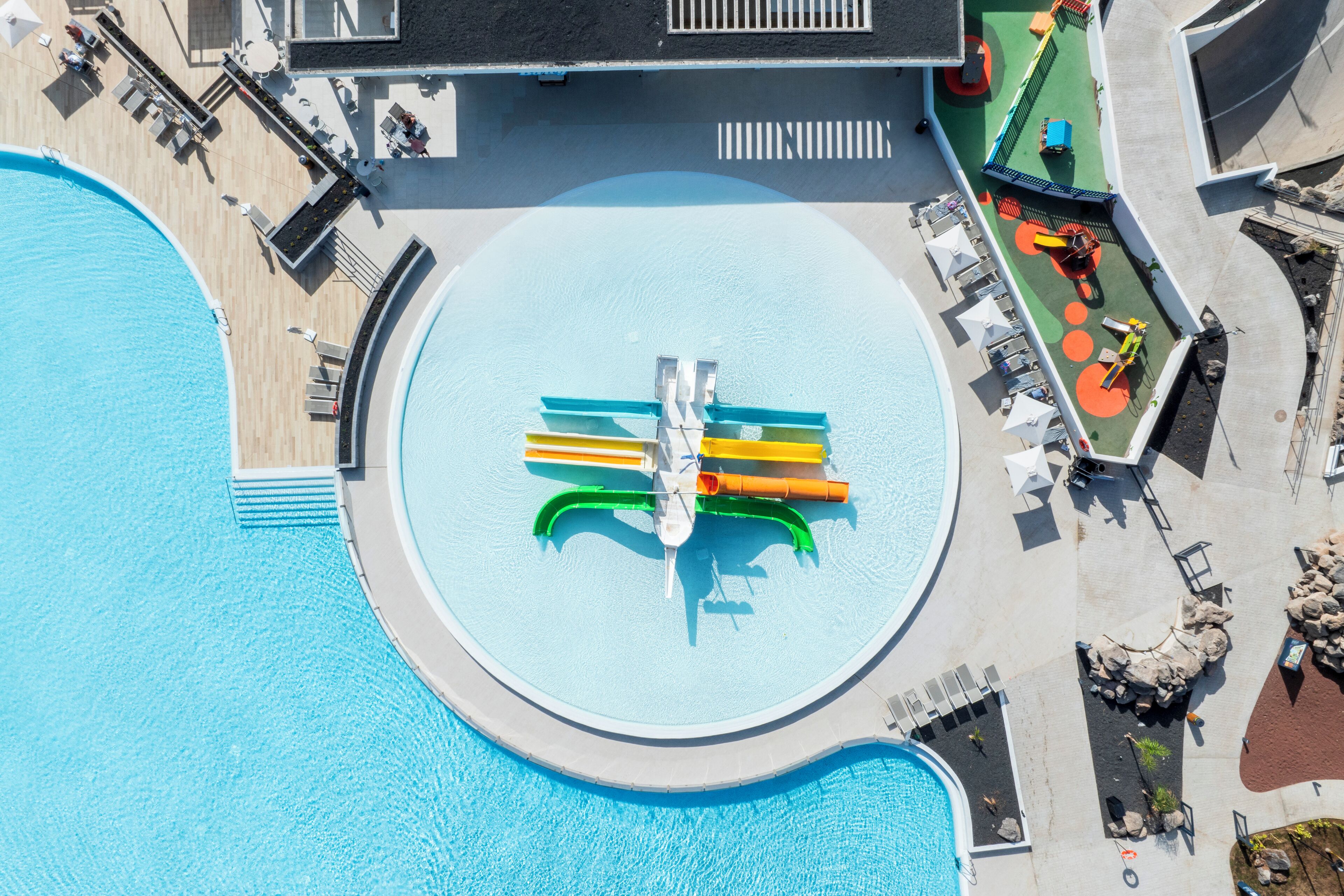 An aerial view of a pool area with a circular water feature, lounge chairs, and a small play area.