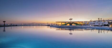 Una piscina infinita tranquila que refleja una puesta de sol, con un puente y sillas de descanso al fondo.