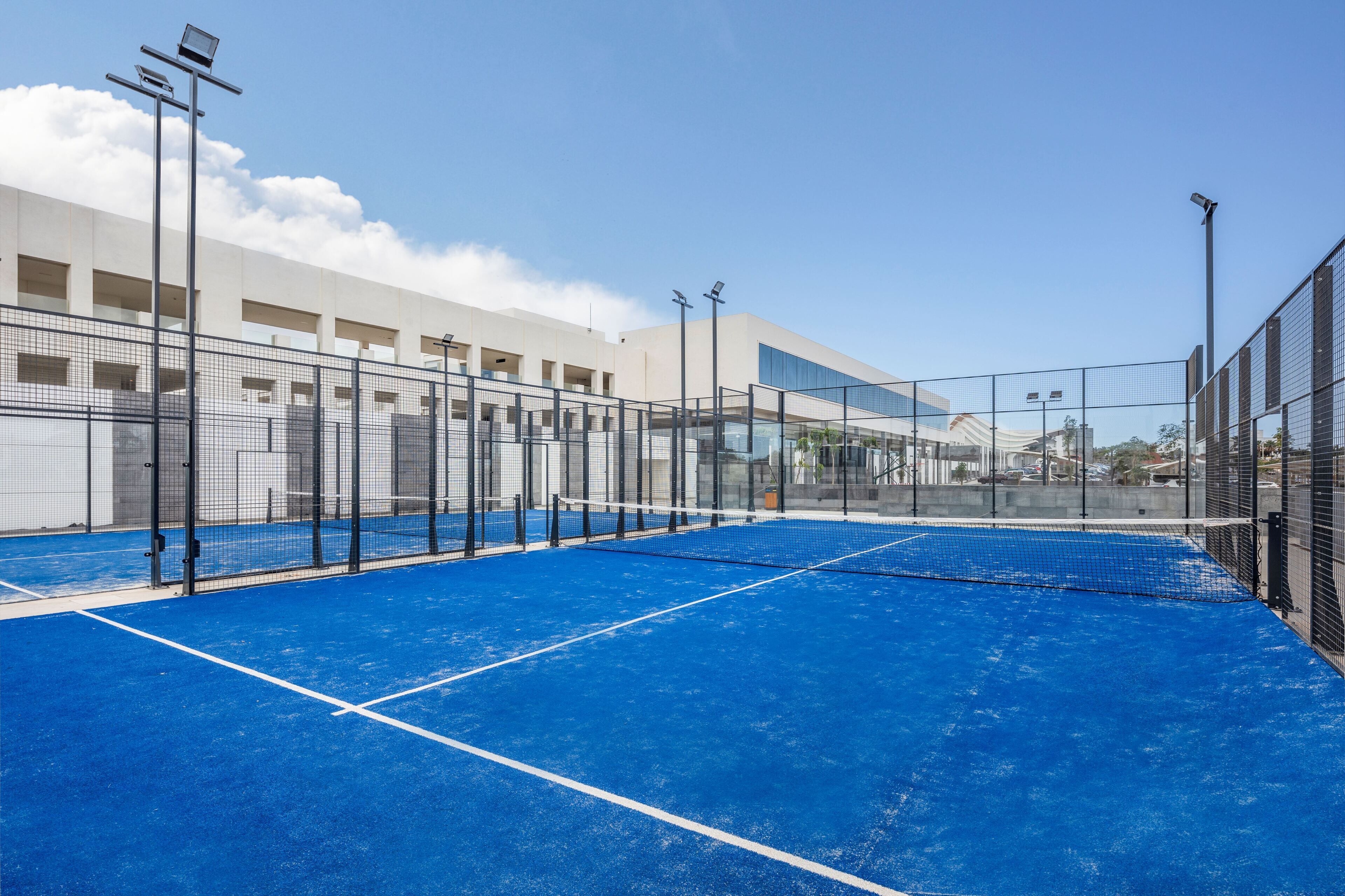 A tennis court with a blue surface and a net, surrounded by a fence, under clear skies.