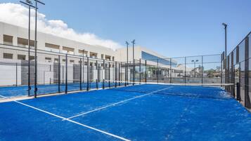 A tennis court with a blue surface and a net, surrounded by a fence, under clear skies.
