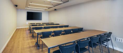 A conference room with a long table, multiple chairs, a flat-screen TV, and ceiling lights.