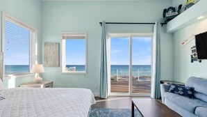 Een slaapkamer met een bed, nachtkastje, lamp, blauw tapijt met een patroon, een grijze bank en uitzicht op het strand door een schuifdeur.