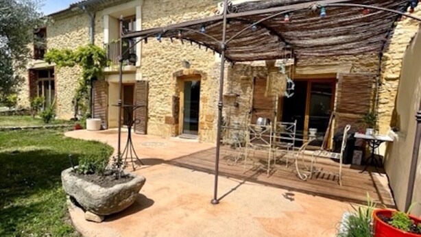 Edificio de piedra con un patio cubierto, asientos exteriores y un jardín con plantas en macetas.