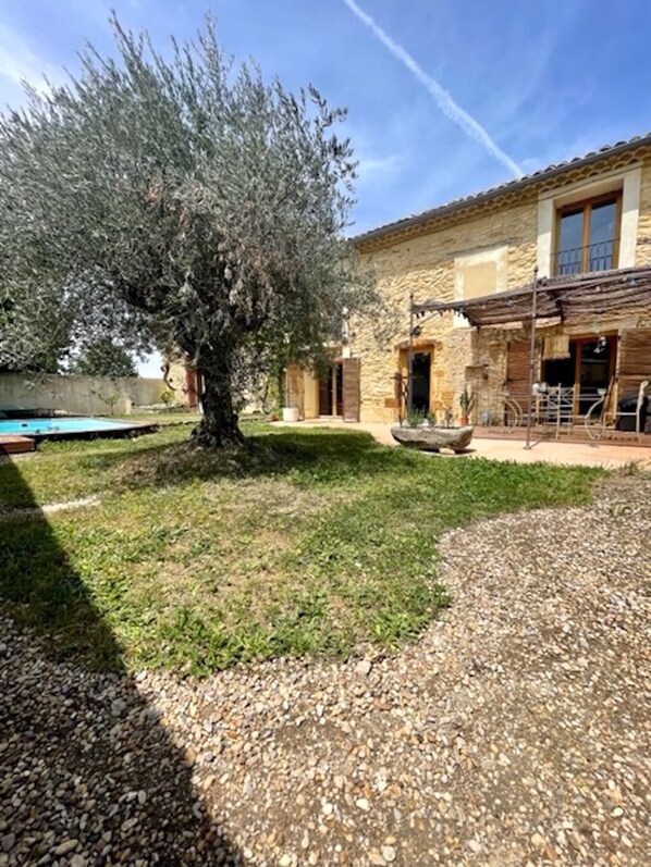 A stone building with a large tree in the yard, a pool, and a gravel area.