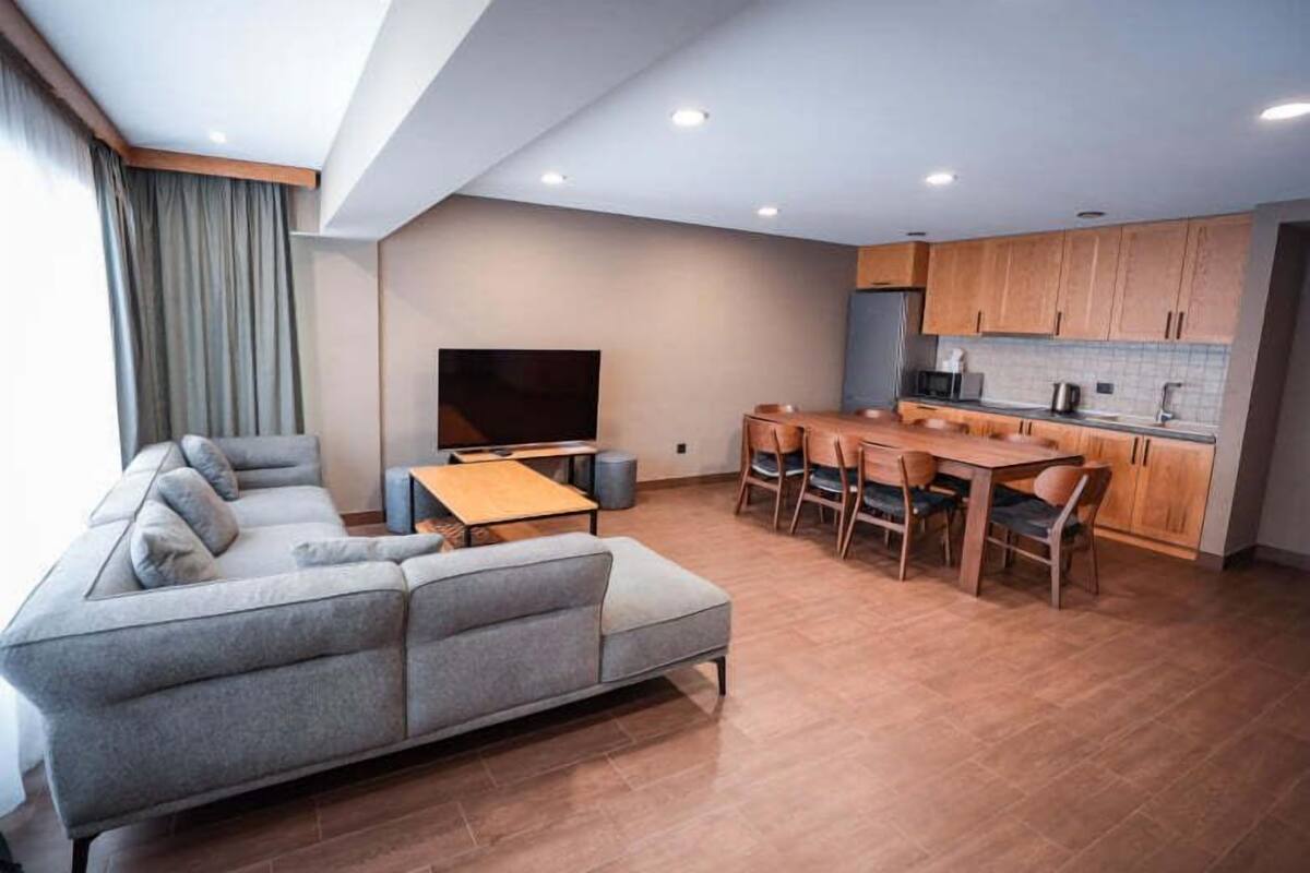 A modern living room with a grey sofa, a wooden dining table, and a kitchen area with wooden cabinets and a flat-screen TV.