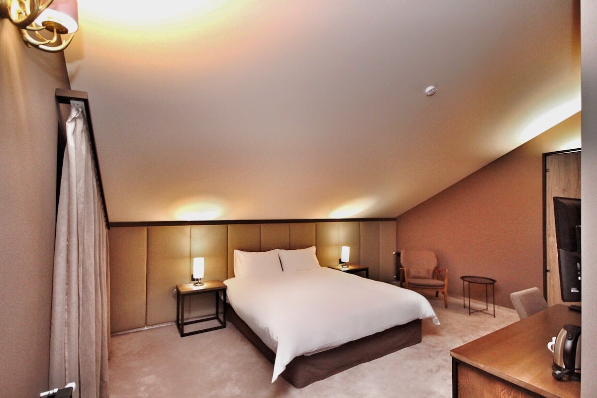 A hotel room with a large bed, bedside tables, a chair, a small table, and a TV.